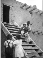 San Ildefonso Pueblo, Corn Dance. War Captain (?) carring youngest participant down steps, two young children watching from bottom